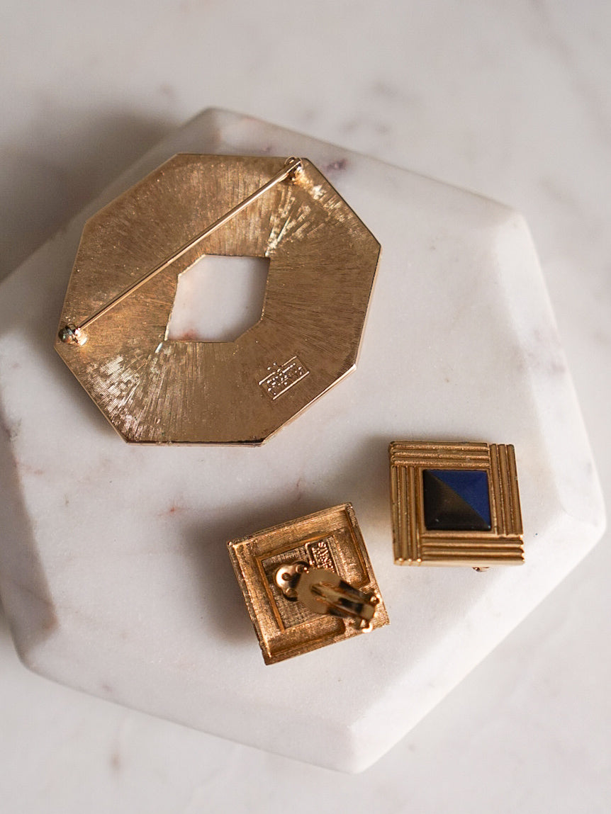 Three vintage gold-toned jewelry pieces, an octagonal brooch with a central cutout, square clip-on earrings with a blue inset.