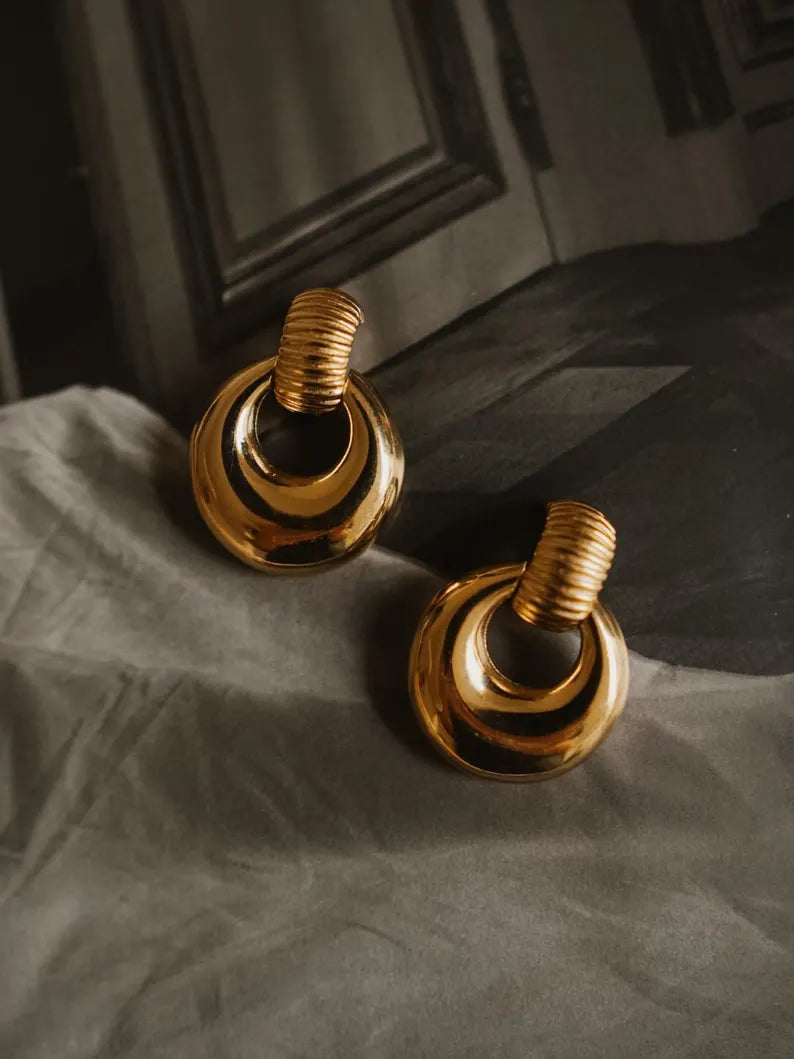 A pair of gold-toned statement earrings with a layered circular design and a textured spiral top post, displayed on draped gray fabric.