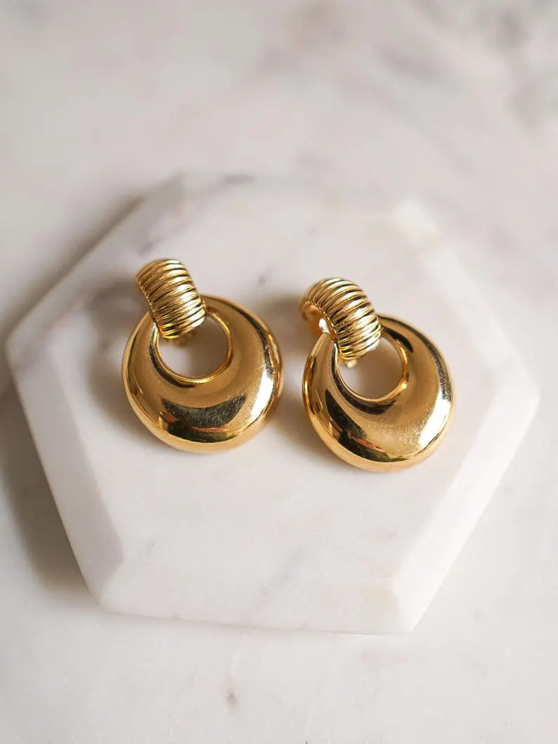 A pair of gold-toned hoop earrings with twisted spiral tops, displayed on a white hexagonal marble stand against a light marble surface.